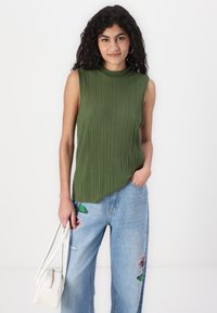 Grønn ermeløs topp med vertikale folder og høy hals, sammen med lyseblå jeans med blomsterbroderi. Hvit håndveske holdt i hånden.