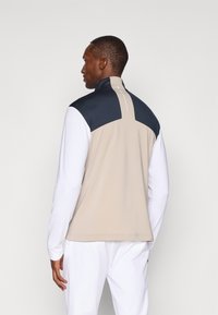 Φωτεινό μπεζ γιλέκο με ναυτικά στοιχεία, που διαθέτει ψηλό κολάρο, μακριά λευκά μανίκια και λεία υφή. Η οπίσθια όψη δείχνει έναν κομψό σχεδιασμό.