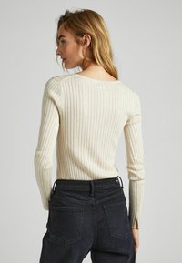 Maglione a coste color crema con maniche lunghe e scollatura ampia, abbinato a jeans scuri con tasche posteriori e dettaglio dell'etichetta.