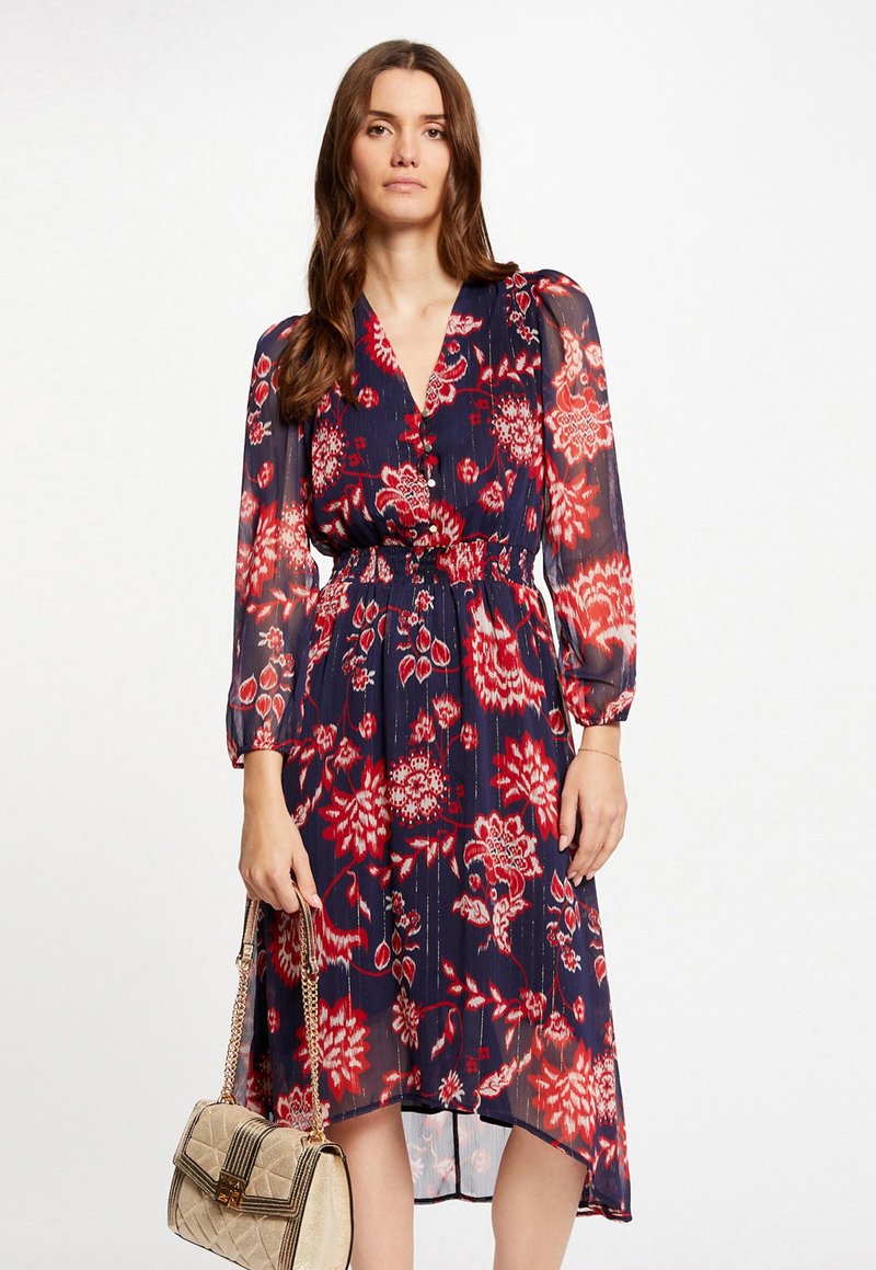 Robe florale marine avec des motifs rouges et blancs, devant boutonné, décolleté en v, et manches longues. Assortie à un sac à main beige à chaîne.