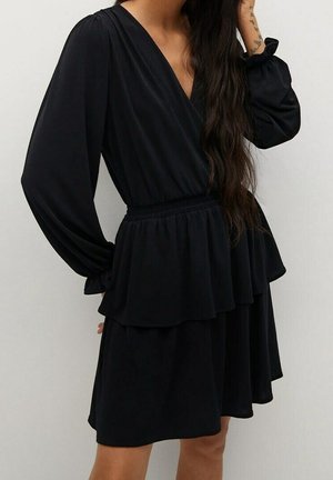 Femme portant une robe noire à manches longues avec un col en V, une taille élastique et une jupe à volants, debout devant un fond uni.