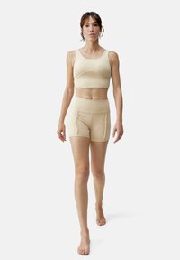 Beige treningsantrekk inkluderer en kort tank topp med brede stropper og høytaljede shorts. Glatt, elastisk stoff med en tettsittende design.
