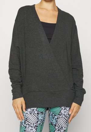Frau trägt dunkelgrauen Langarm-V-Ausschnitt-Pullover über einem schwarzen Top mit gemusterten Leggings mit grünen, weißen und blauen Blumen- und Streifendekoren.