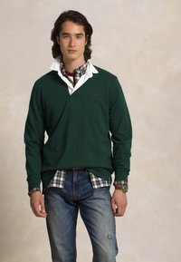 Grønn langermet polo skjorte lagdelt over en hvit skjorte med krage med rutemønster, brukt sammen med blå jeans. Merkelig logo på brystet.