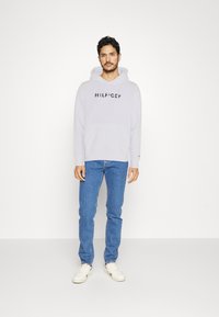 Felpa grigia chiara con tasca frontale e logo centrato sul petto, abbinata a jeans blu e sneakers bianche. Design minimalista, vestibilità rilassata.