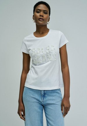 Joven mujer con el pelo corto que lleva una camiseta blanca "SALSA JEANS" y vaqueros azul claro, de pie frente a un fondo gris liso.