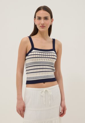 Giovane donna con capelli castani lisci che indossa un top senza maniche in maglia blu navy e bianco e una gonna bianca con coulisse, in piedi davanti a uno sfondo semplice.