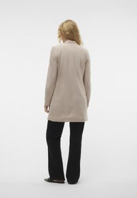 Abrigo largo beige de corte entallado, con mangas largas, cuello y una textura suave, combinado con pantalones de campana negros y zapatos planos.