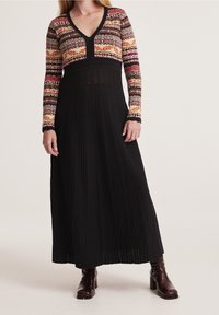 Maxi šaty s barevným vzorovaným vrškem a dlouhou černou plisovanou sukní. Mají výstřih do V a dlouhé rukávy; textura pletené tkaniny.