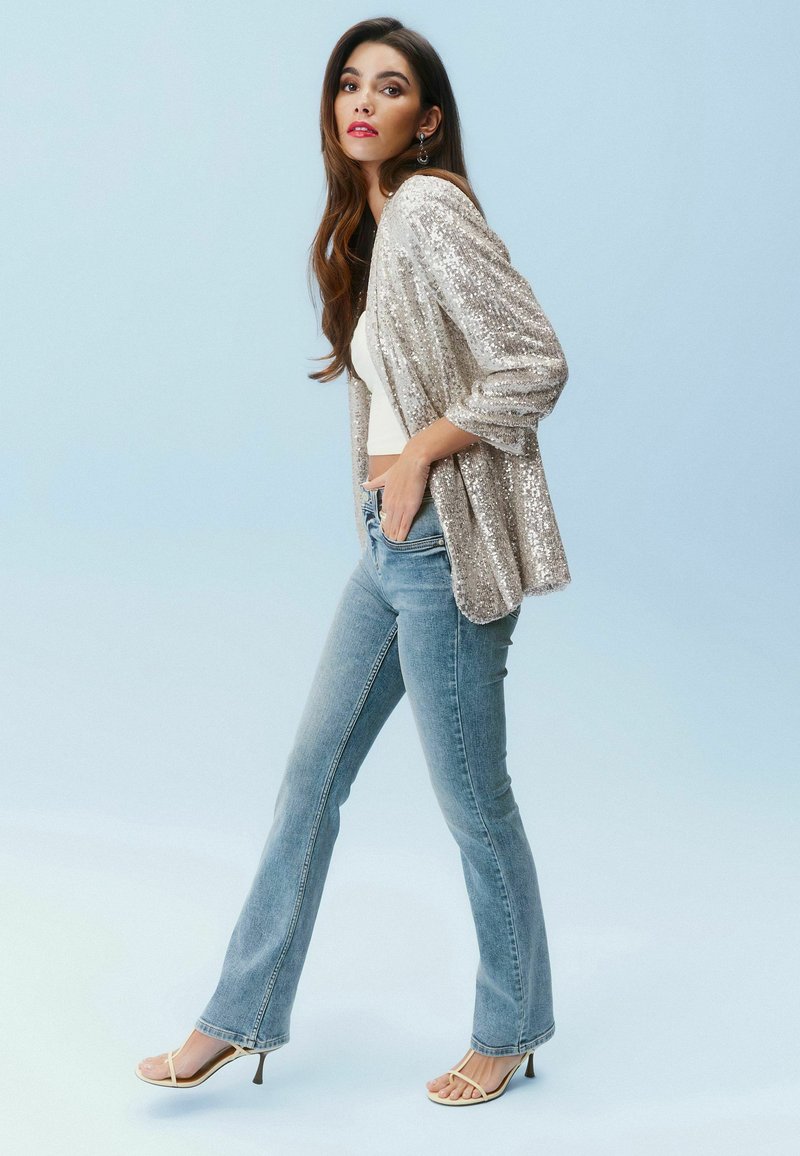 Helles graues Pailletten-Blazer über einem weißen Crop-Top, kombiniert mit blauen Schlagjeans und Riemchenheels. Das Model posiert vor einem sanften blauen Hintergrund.