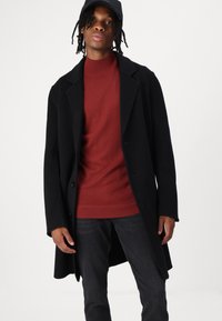 Cappotto di lana nero sopra un dolcevita rosso a costine, jeans scuri e berretto nero. Il cappotto presenta un colletto a risvolto e chiusura con bottoni.
