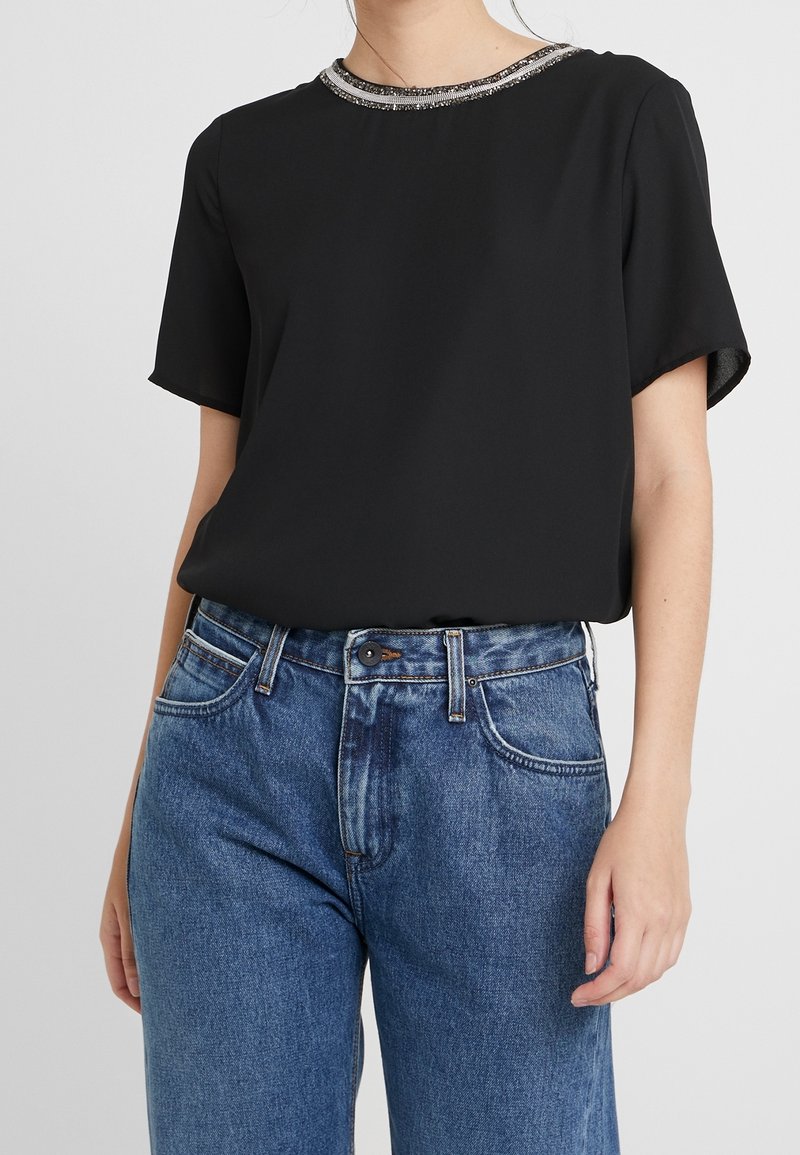 Top negro de manga corta con un cuello de cuentas, hecho de tela ligera. Combinado con jeans de denim azul de cintura alta.