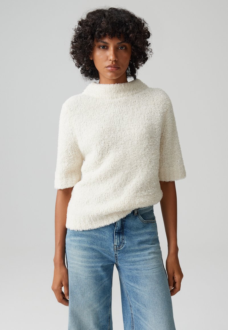 Maglione a maglia con trama, di colore crema, con colletto alto e maniche corte, abbinato a jeans a gamba larga di colore blu chiaro. Tessuto morbido, vestibilità comoda.