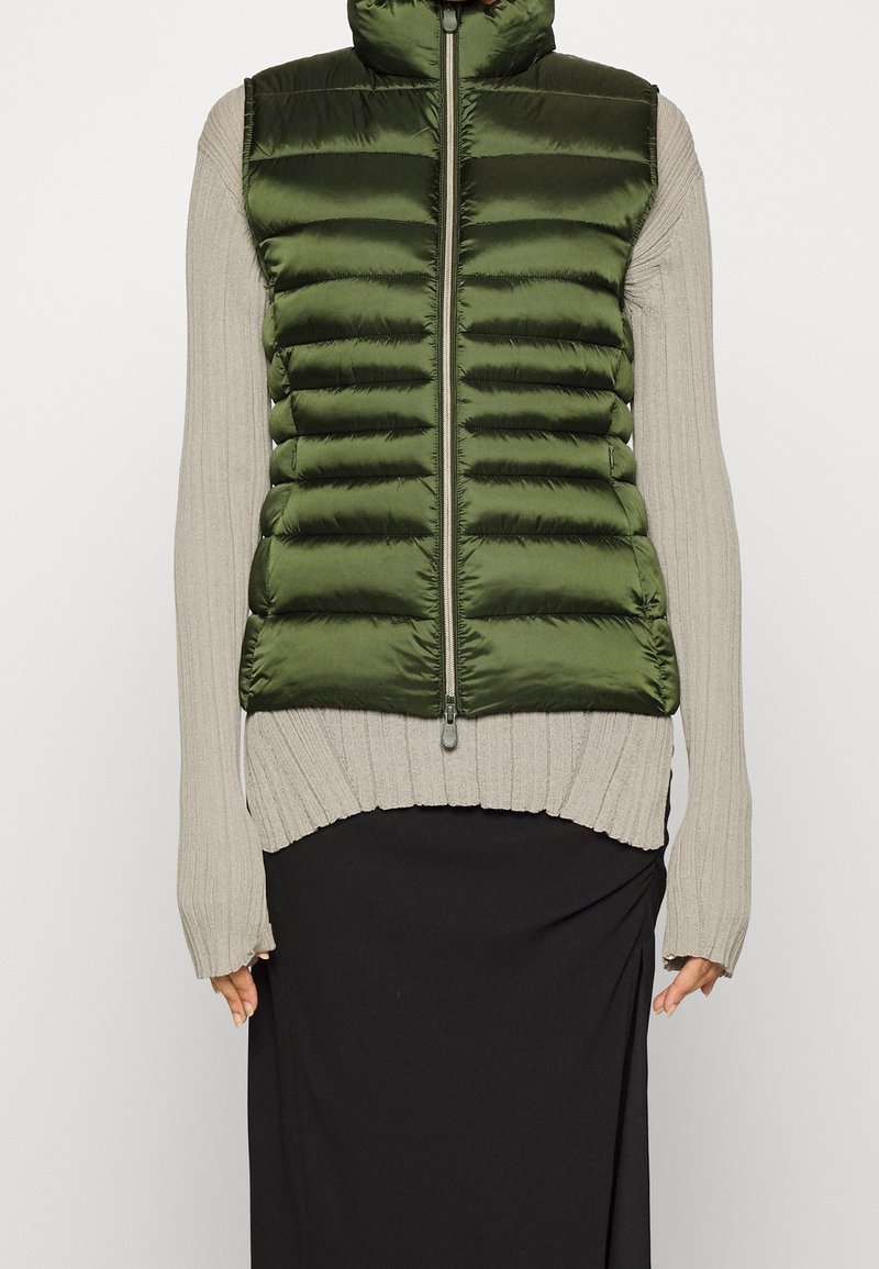 Grøn polstret vest med høj krave, lynlåsfortil og quiltet design, over en ribbet langærmet bluse, parret med en sort nederdel.