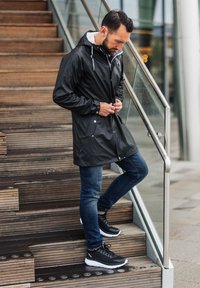 Sort vandtæt jakke med hætte, der har trykknapper og to frontlommer. Bæres over blå jeans og sorte sportsko med hvide såler.