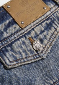 Ύφασμα τζιν με μπλε και ανοιχτό μπλε σχέδιο, δερμάτινο έμπλαστρο με την ετικέτα «Juicy Couture Jeans» και ασημένια μεταλλικά στοιχεία με λεπτομέρεια λογότυπου.