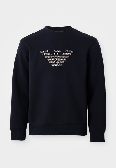 Tamsiai mėlynas džemperis su ilgomis rankovėmis, su raštuota erelio logotipu ir „GA“ inicialais, išdėstytais viduryje krūtinės.