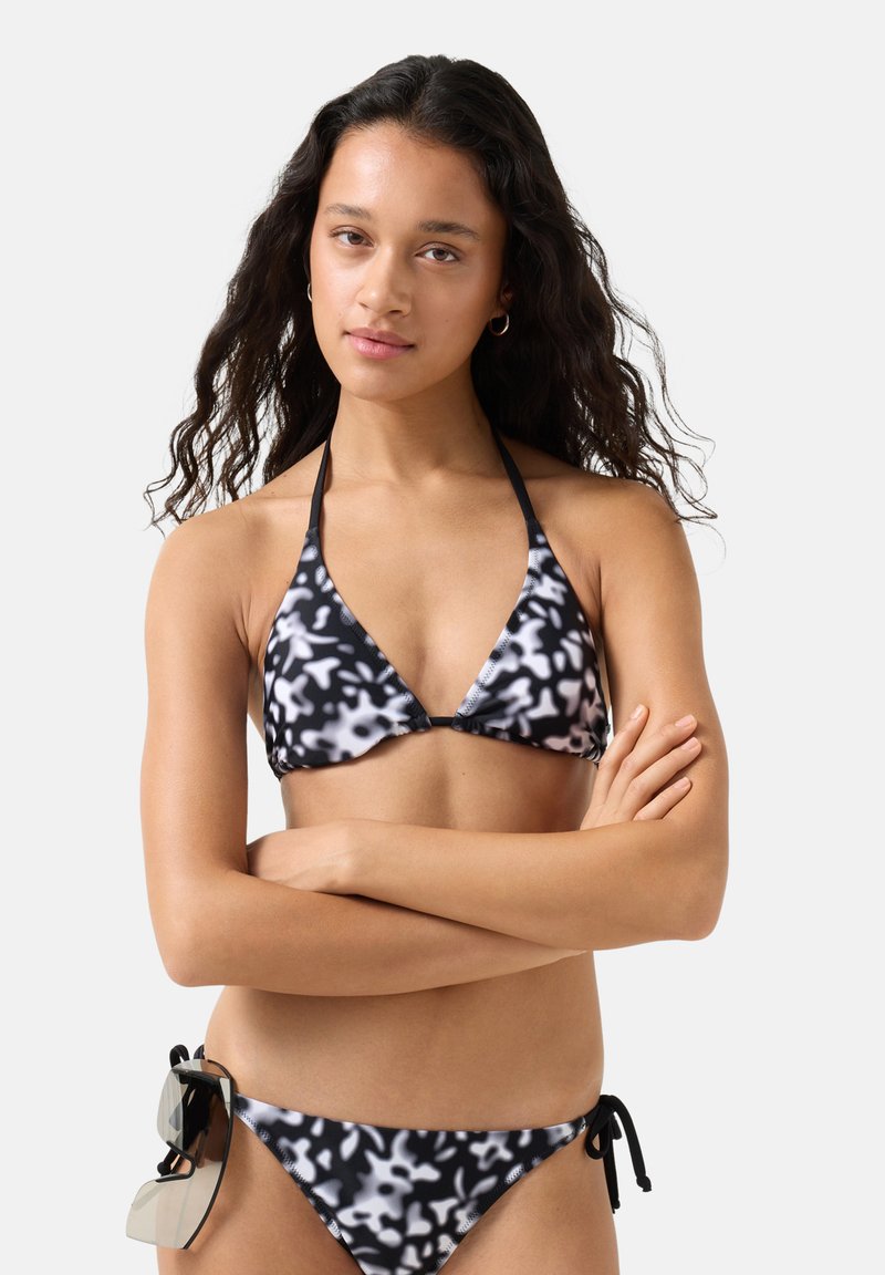 Frau mit langem, dunklem Haar, die einen schwarz-weiß gemusterten Bikini mit seitlichen Schnürungen trägt und eine Sonnenbrille an der Hüfte hält, die Arme verschränkt.