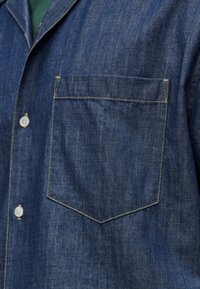 Devant d'une veste en denim avec une poche poitrine visible, des coutures jaunes, des boutons blancs, et un aperçu d'un vêtement vert en dessous.