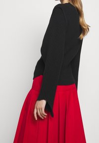 Schwarzer gerippter Pullover mit langen, ausgestellten Ärmeln, kombiniert mit einem fließenden roten plissierten Rock. Die Stoffstruktur ist auf beiden Kleidungsstücken sichtbar.