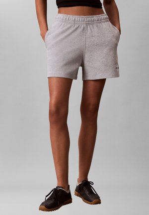 Persoon draagt grijze shorts met elastische taille en zakken, een zwart cropped top en zwarte sneakers met bruine zolen, staand tegen een grijze achtergrond.