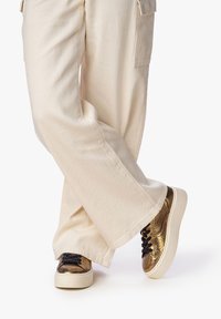 Sneakers metallici dorati con una finitura texturizzata, lacci neri e pantaloni in tessuto color crema. I sneakers presentano una suola bianca chunky.