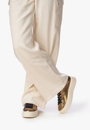 Sneakers metallici dorati con una finitura texturizzata, lacci neri e pantaloni in tessuto color crema. I sneakers presentano una suola bianca chunky.