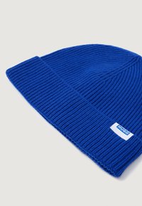 Căciulă din tricot ribat albastru, cu tiv pliant și o etichetă albă pe care este scris "HUGO." Material texturat și ajustare strânsă.