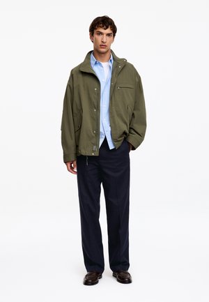 Jeune homme portant une veste vert olive sur une chemise bleu clair, un pantalon foncé et des chaussures marron foncé, debout avec une main dans la poche.