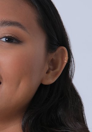 Boucle d'oreille en or exposée sur l'oreille d'un modèle. Le design présente une apparence torsadée et superposée et est installée sur le cartilage.