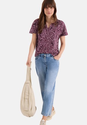 Femme portant un haut à manches courtes violet à motif cachemire, un jean bleu clair, des baskets beiges, tenant un sac à bandoulière beige matelassé dans une main.