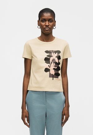 Lyhytukkainen nainen pukeutuneena beigeen T-paitaan, jossa on vaaleanpunainen ja musta kukkakuvio, sekä vaaleansinisiin housuihin, seisoo yksivärisen taustan edessä.