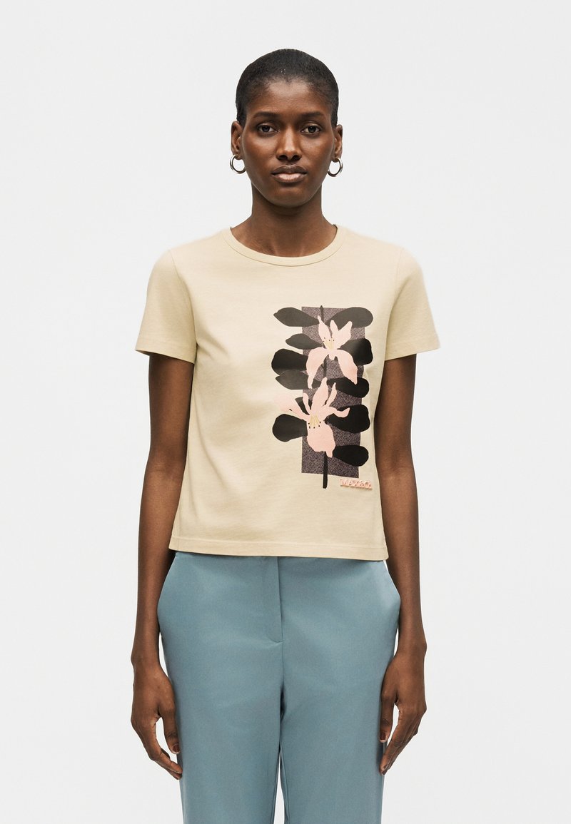 Γυναίκα με κοντά μαλλιά φοράει μπεζ μπλουζάκι με ροζ και μαύρο λουλουδάτο σχέδιο και ανοιχτό μπλε παντελόνι, στέκεται μπροστά σε ένα απλό φόντο.