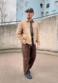 Light beige cotton jacket, patterned black shirt, brown pants, and black sneakers. Includes chain detail on pants and a dark cap.