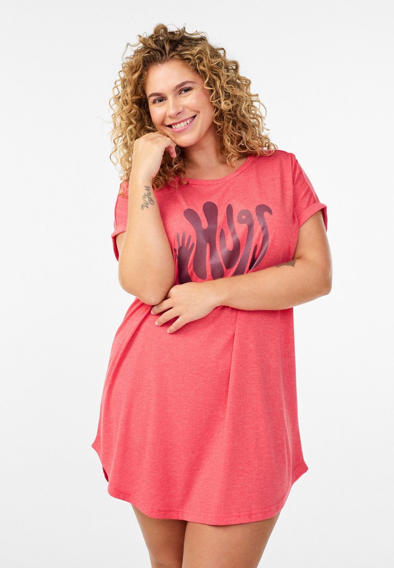 Lächelnde Frau mit lockigem blondem Haar trägt ein korallenfarbenes Kleid mit dem Schriftzug "Hugs" und einem Handabdruck-Design, posiert mit der Hand am Kinn vor weißem Hintergrund.