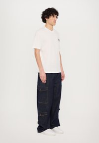 Camiseta de algodón blanca con mangas cortas, detalle gráfico negro en el pecho, combinada con pantalones cargo de pierna ancha en azul oscuro con múltiples bolsillos.
