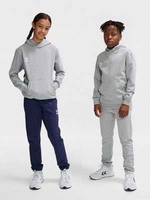 Dos niños están de pie vistiendo sudaderas con capucha grises a juego y zapatillas blancas, uno con pantalones azul marino y el otro con pantalones deportivos grises.