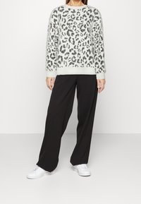 Leopardenmuster-Pullover in Hellgrau mit schwarzen Akzenten, kombiniert mit schwarzen weiten Hosen und weißen Sneakers. Glatte Textur, entspannter Schnitt.