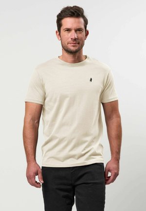 Homme aux cheveux bruns et courts, portant un t-shirt beige à manches courtes et un pantalon noir, debout devant un fond blanc uni.