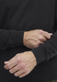 Pull en maille noire avec poignets côtelés, tissu subtilement texturé et coupe ajustée, montrant des mains ajustant la manche.