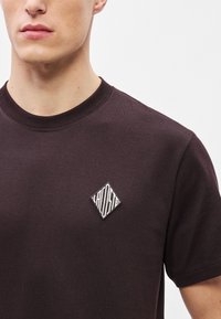 Ciemnobrązowa bawełniana koszulka z okrągłym dekoltem i teksturowaną naszywką z logo w kształcie diamentu w kolorze białym i czarnym na lewym przednim panelu.