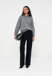 Szürke galléros pulóver, tengerészkék, széles lábú nadrág, fekete clutch tartása. Lapos fekete cipők. Göndör haj, egy sima háttér előtt áll.