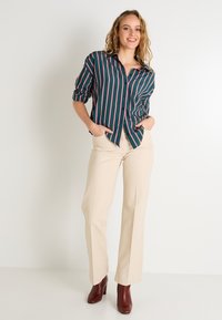 Camisa de botones a rayas en verde oscuro con líneas rosas; combinada con pantalones beige; modelo llevando botines de tacón burdeos.