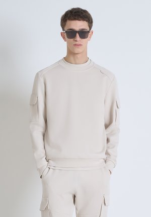 Beige crew-neck sweatshirt with ribbed collar, side seams, and a pocket on the left sleeve. Smooth texture and relaxed fit.
