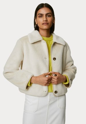 Cremefarvet falsk pelsjakke med krave og to brune knapper, båret over en struktureret lys gul sweater. Glat og hyggelig tekstur.