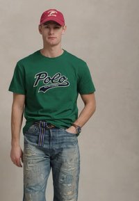 Πράσινη βαμβακερή μπλούζα με λογότυπο "Polo Ralph Lauren", σε συνδυασμό με φθαρμένα μπλε τζιν. Κόκκινο καπέλο και πλεκτή ζώνη με πολύχρωμες ρίγες.