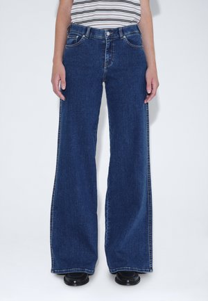 Persoon draagt donkerblauwe high-waist wijde jeans met een gestreept mouwloos topje en zwarte schoenen, staand tegen een effen witte achtergrond.