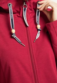 Sweat à capuche rouge zippé avec cordons de serrage bleu marine et blanc ornés de perles en bois et texte de marque, main ajustant un des cordons.