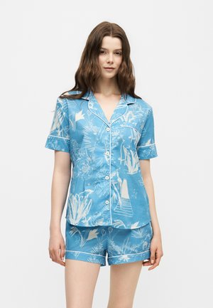 Tânără purtând o cămașă albastră cu mâneci scurte cu nasturi și pantaloni scurți asortați cu modele botanice albe pe un fundal simplu.