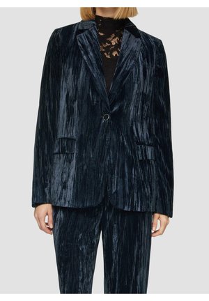 Femme portant un blazer en velours froissé bleu foncé et un pantalon assorti sur un top en dentelle noire, debout devant un fond clair uni.